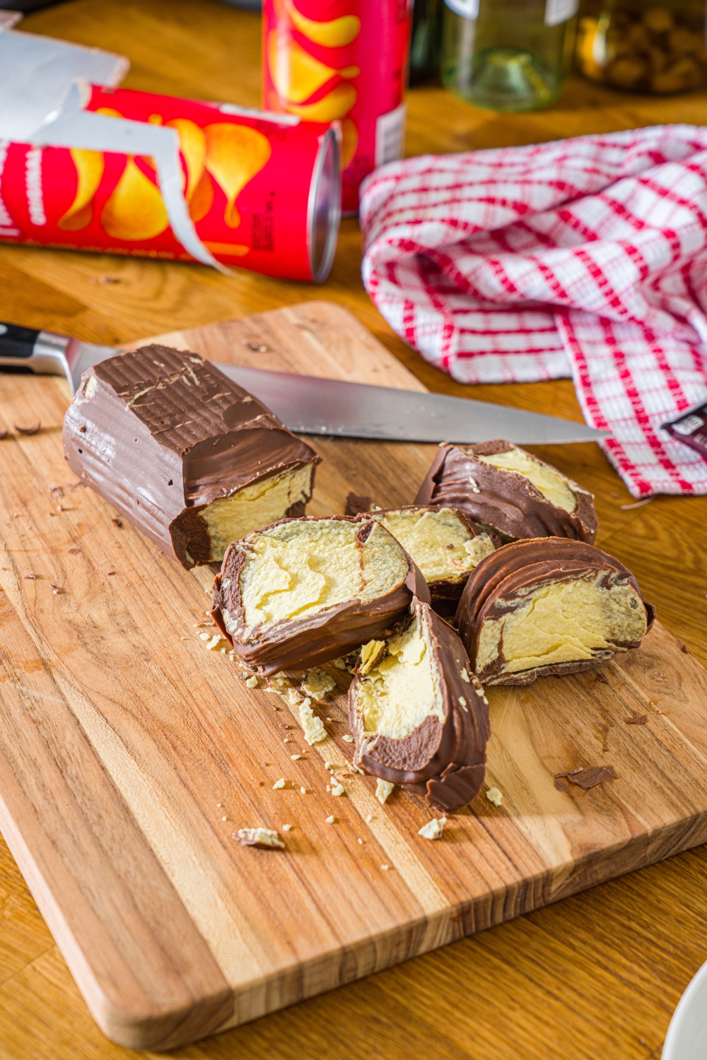 A few pieces of chocolate covered Pringles in front of the Pringles chocolate black all on a wooden cutting board.