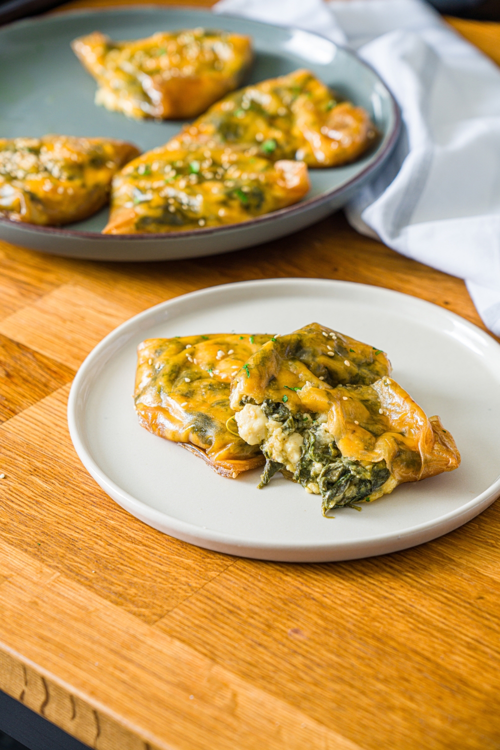 A white plate with rice paper spanakopita split open. The plate is on a wooden counter with an additional plate of spanakopita and a white cloth napkin.