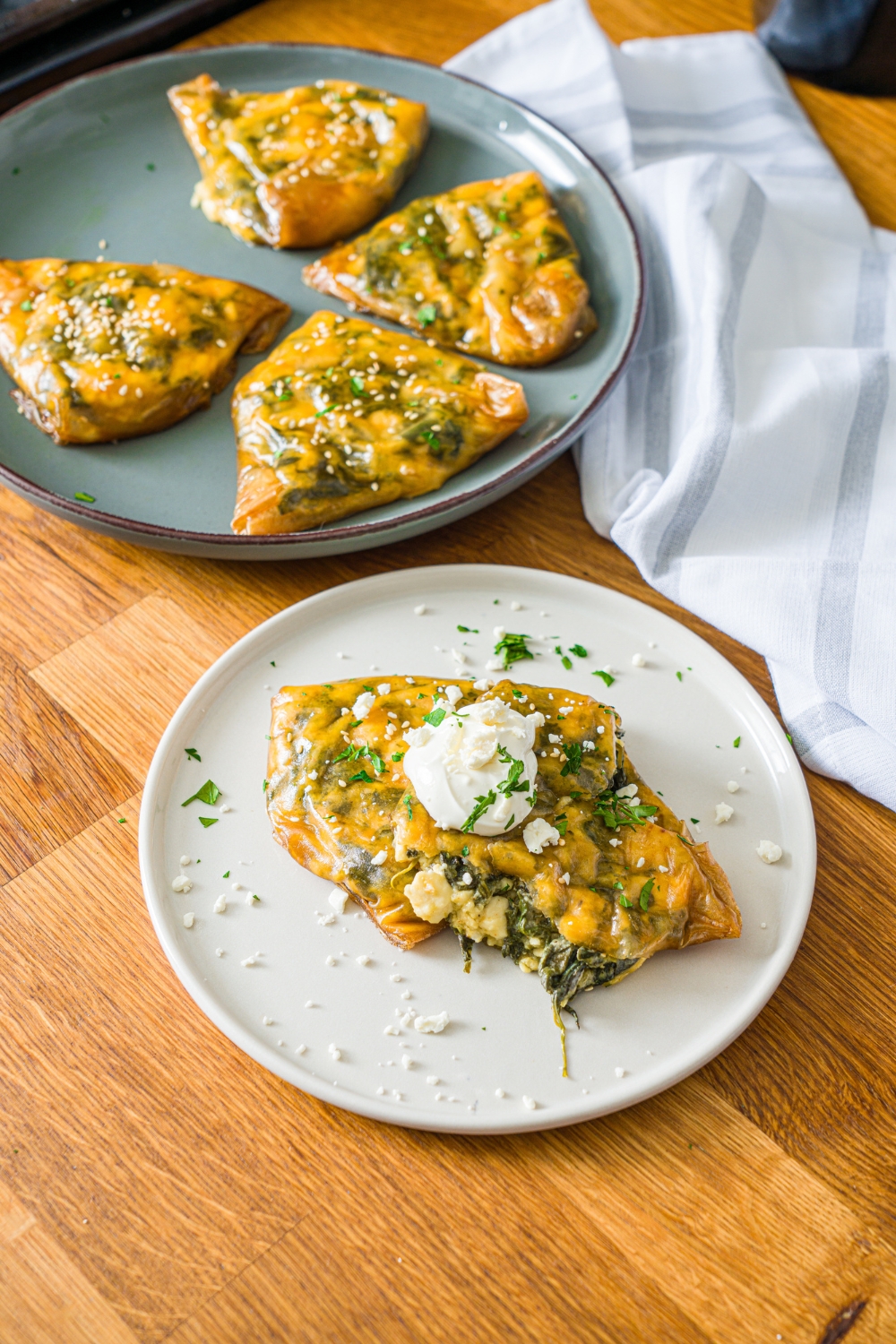 A white plate with rice paper spanakopita split open and topped with sour cream. The plate is garnished with feta cheese and fresh parsley. The plate is on a wooden counter with an additional plate of spanakopita and a white cloth napkin.