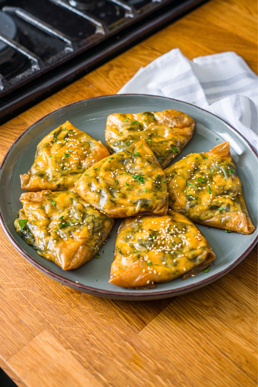 A green plate with several pieces of rice paper spanakopita finished with sesame seeds and fresh parsley. The plate is on a wooden counter with a white cloth napkin.
