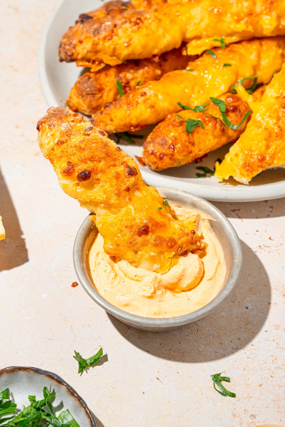 A cottage cheese breadstick dipped in a small cup of dipping sauce. The cup is on a tan counter with a plate of breadsticks.