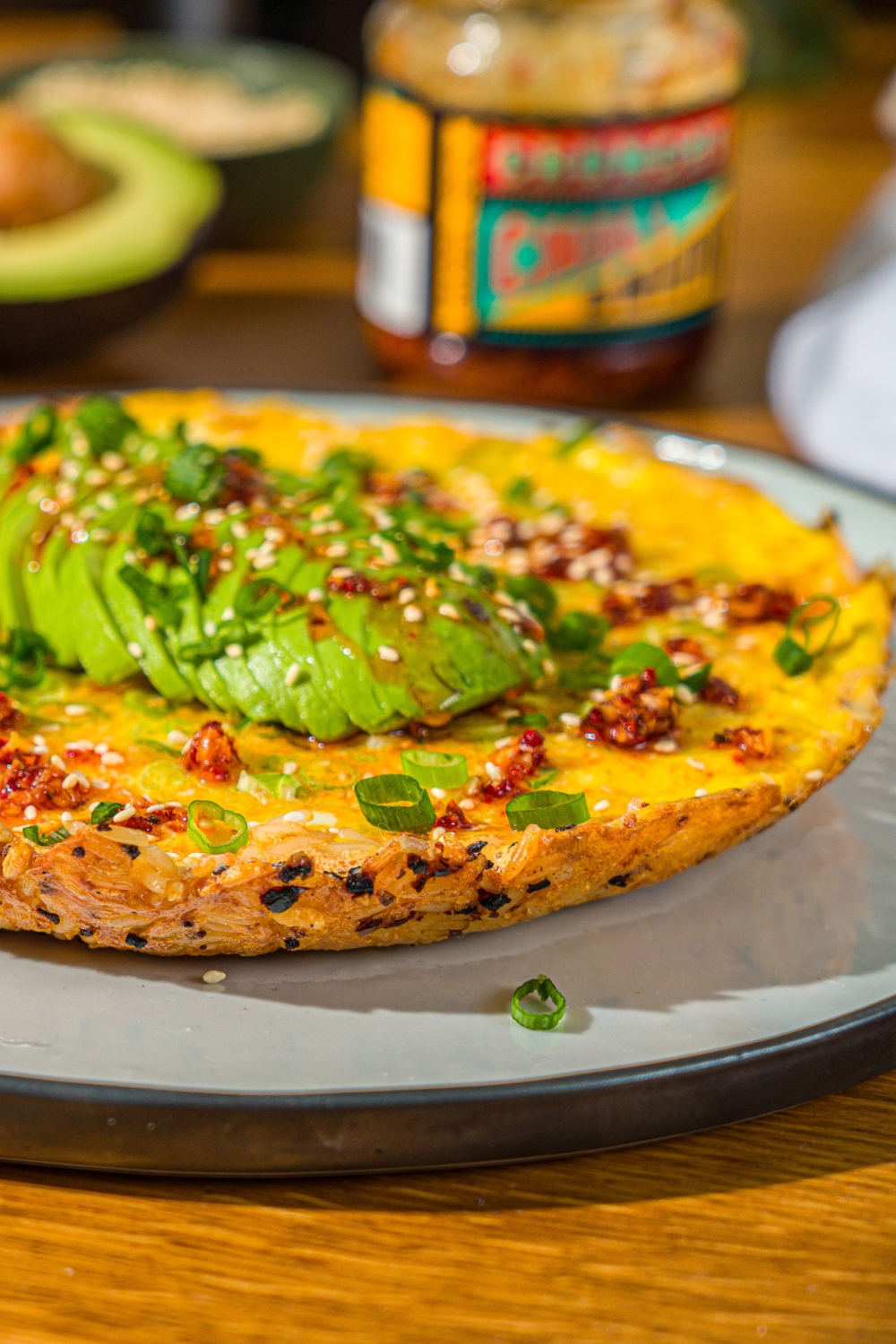 A white plate with a crispy rice omelette topped with sliced avocado, chili crunch oil, and sliced green onion. The plate is on a wooden counter with small bowls of garnishes and an avocado.