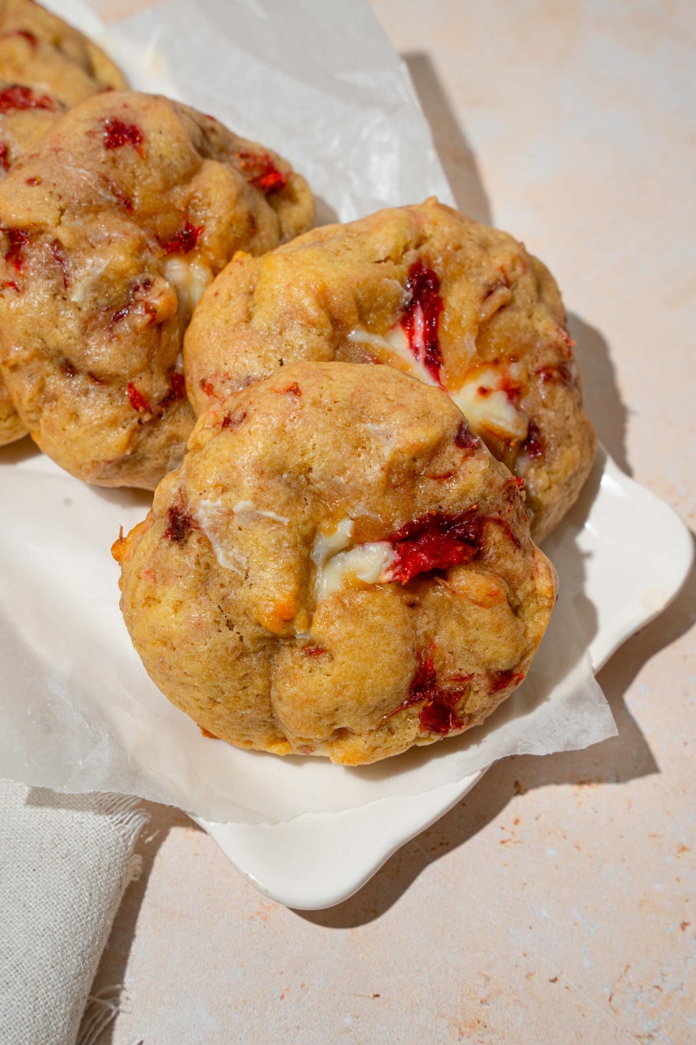 A white plate lined with parchment paper with several strawberry cheesecake cookies. The plate is on a tan counter.