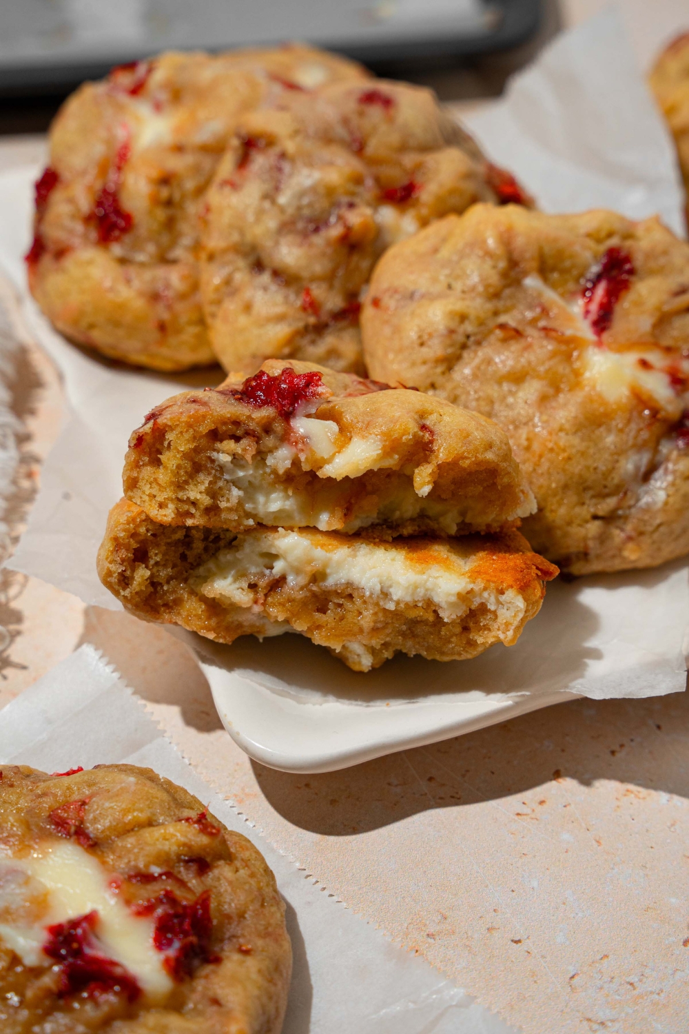 A white plate lined with parchment paper with several strawberry cheesecake cookies. One of the cookies is sliced in half and stacked on one another. The plate is on a tan counter.