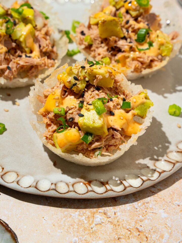 A plate with three tuna rice cups topped with avocado, sliced green onion, sesame seeds, and spicy mayo. The plate is on a tan counter.