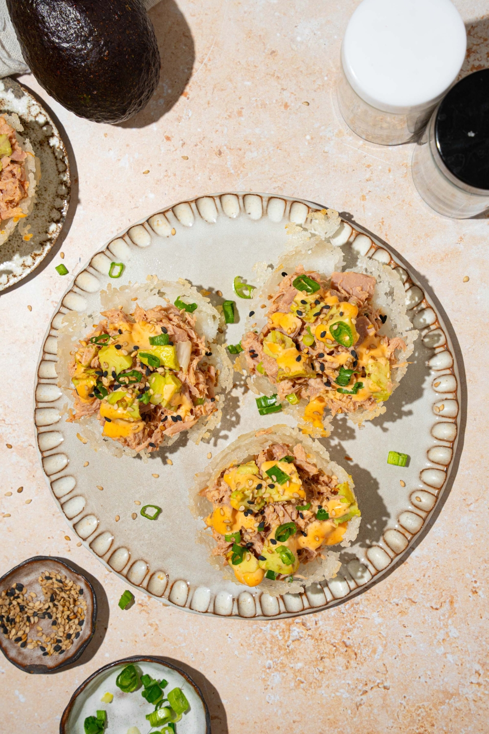 A plate with three tuna rice cups topped with avocado, sliced green onion, sesame seeds, and spicy mayo. The plate is on a tan counter with small plates of garnishes.