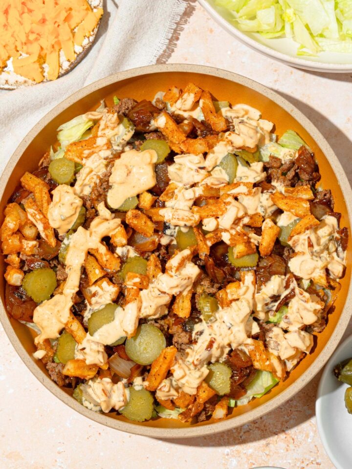 A burger bowl with burger sauce, ground beef, fries, lettuce, pickles, caramelized onions, and cheese in a yellow bowl on top of a white counter.