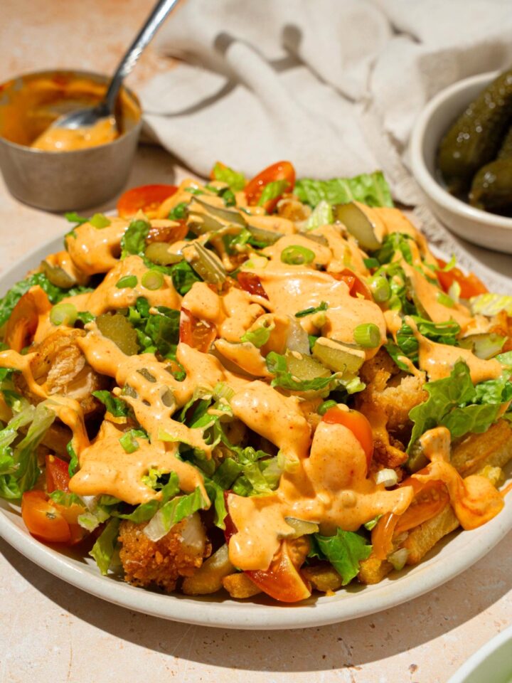 Chicken burger bowl loaded fries on a white plate with crispy chicken, french fries, lettuce, tomatoes, melted cheese, and a burger sauce on top.