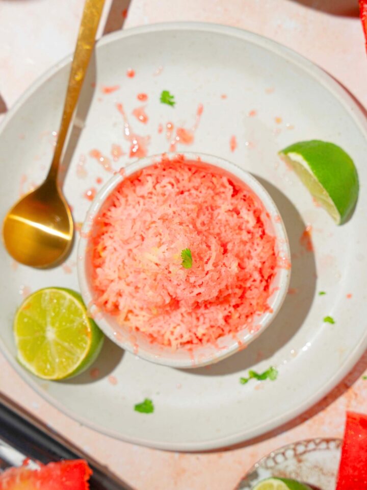 Watermelon shaved ice in a white cup on top of a white plate with a gold spoon and a lime wedge on it.