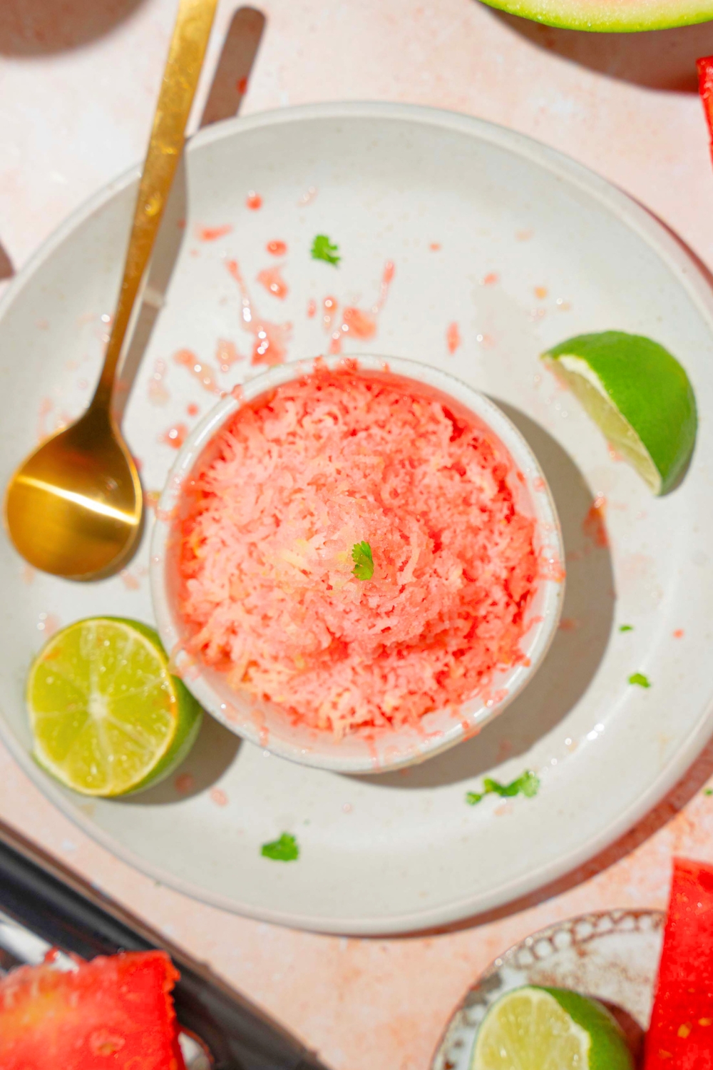 Watermelon shaved ice in a white cup on top of a white plate with a gold spoon and a lime wedge on it.