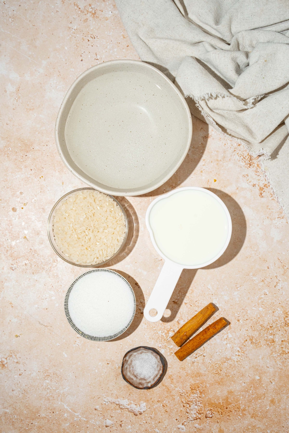 A tan counter with several bowls in various sizes containing ingredients to make arroz con leche including rice, water, cinnamon sticks, milk, sugar, and salt.