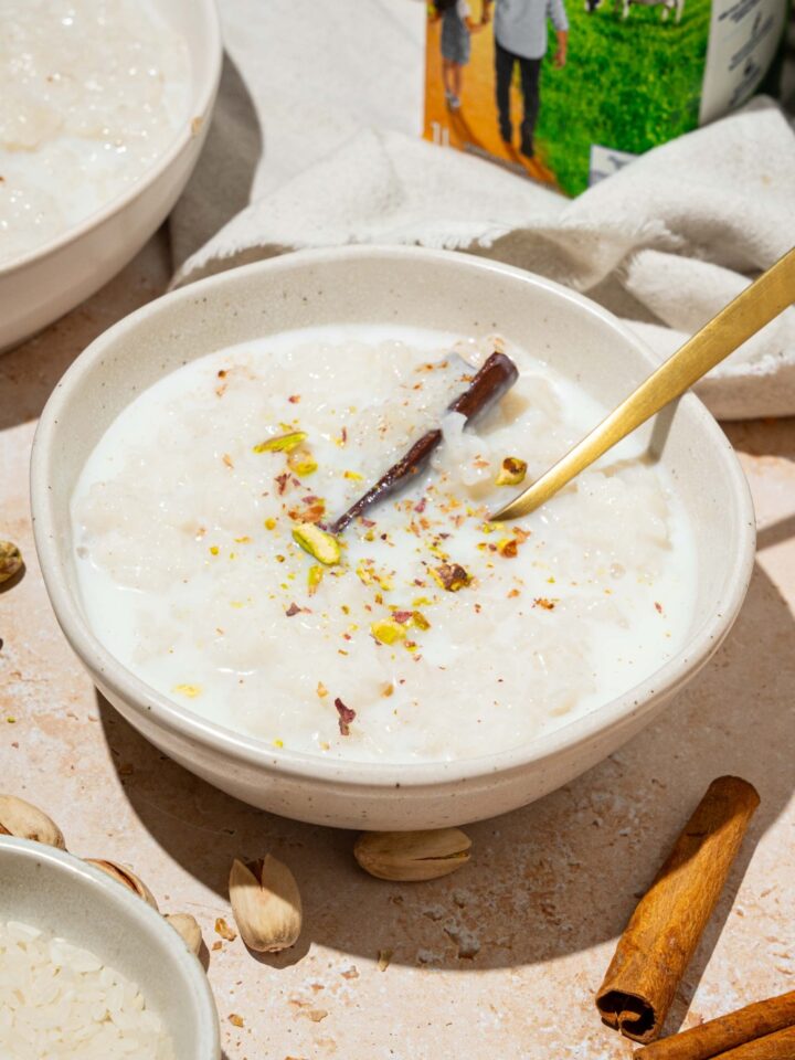 A white bowl with arroz con leche topped with a cinnamon stick and pistachios. There is a spoon in the bowl. The bowl is on a tan counter with an additional bowl of rice and a white cloth napkin.