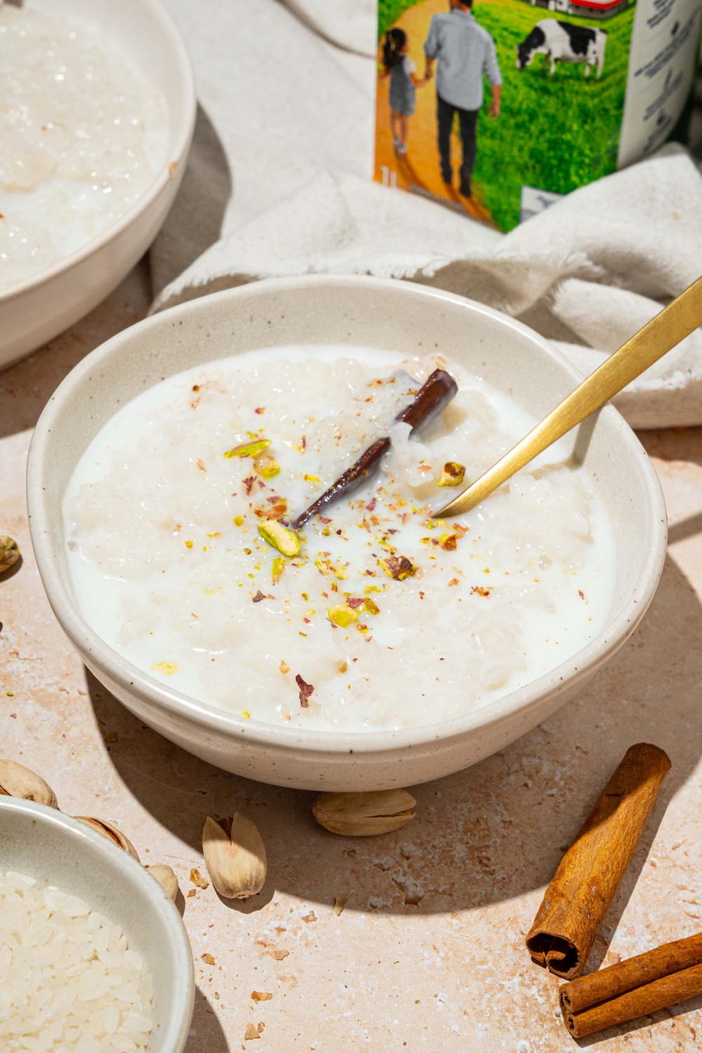 A white bowl with arroz con leche topped with a cinnamon stick and pistachios. There is a spoon in the bowl. The bowl is on a tan counter with an additional bowl of rice and a white cloth napkin.