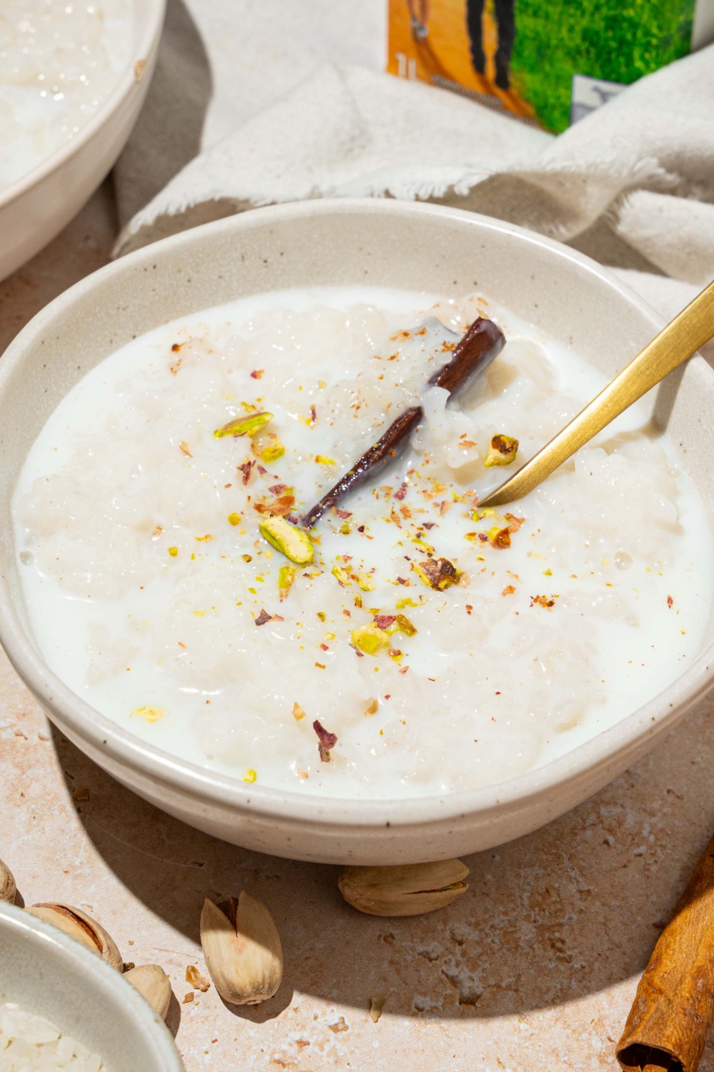 A white bowl with arroz con leche topped with a cinnamon stick and pistachios. There is a spoon in the bowl. The bowl is on a tan counter with an additional bowl of rice and a white cloth napkin.