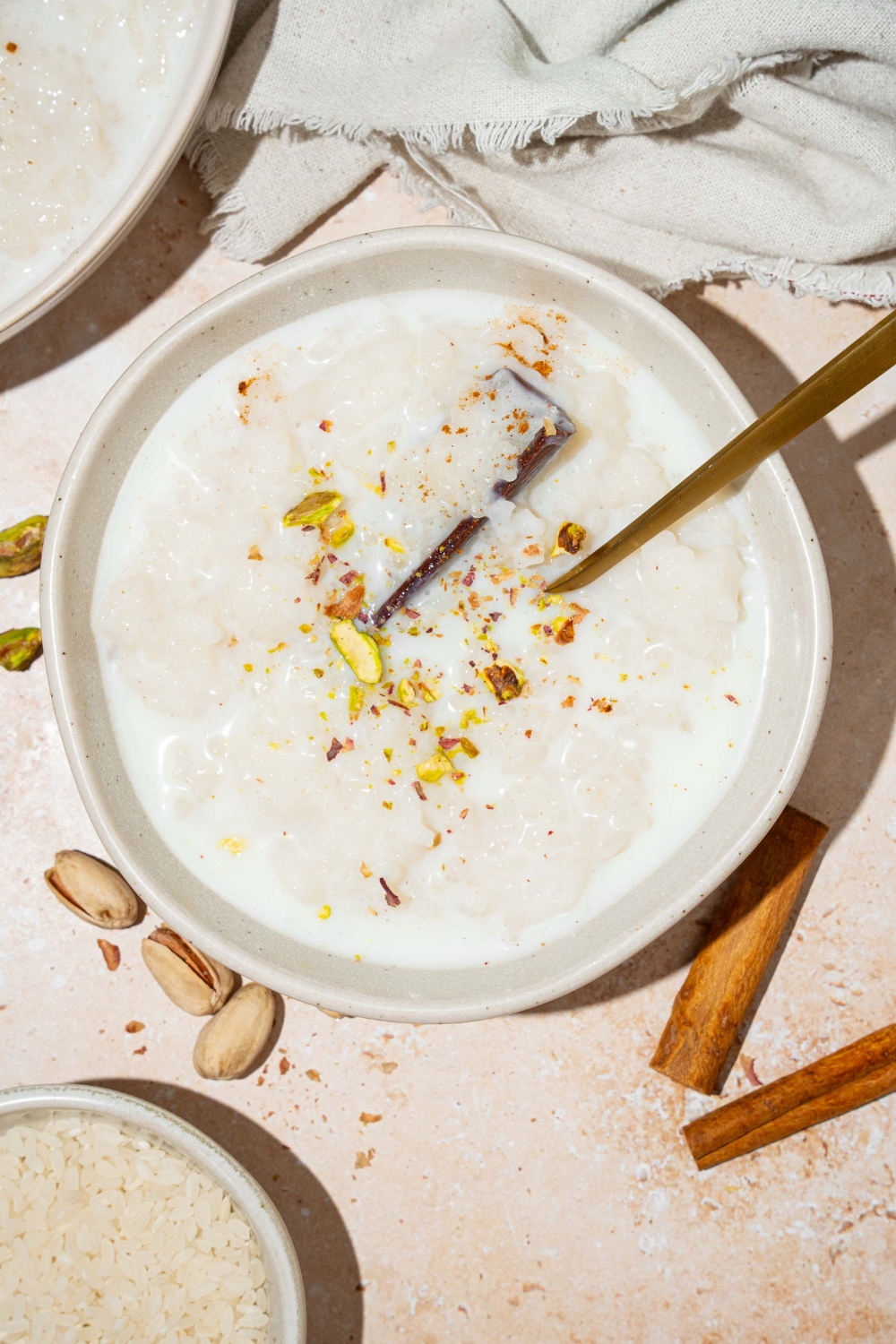 A white bowl with arroz con leche topped with a cinnamon stick and pistachios. There is a spoon in the bowl. The bowl is on a tan counter with an additional bowl of rice and a white cloth napkin.