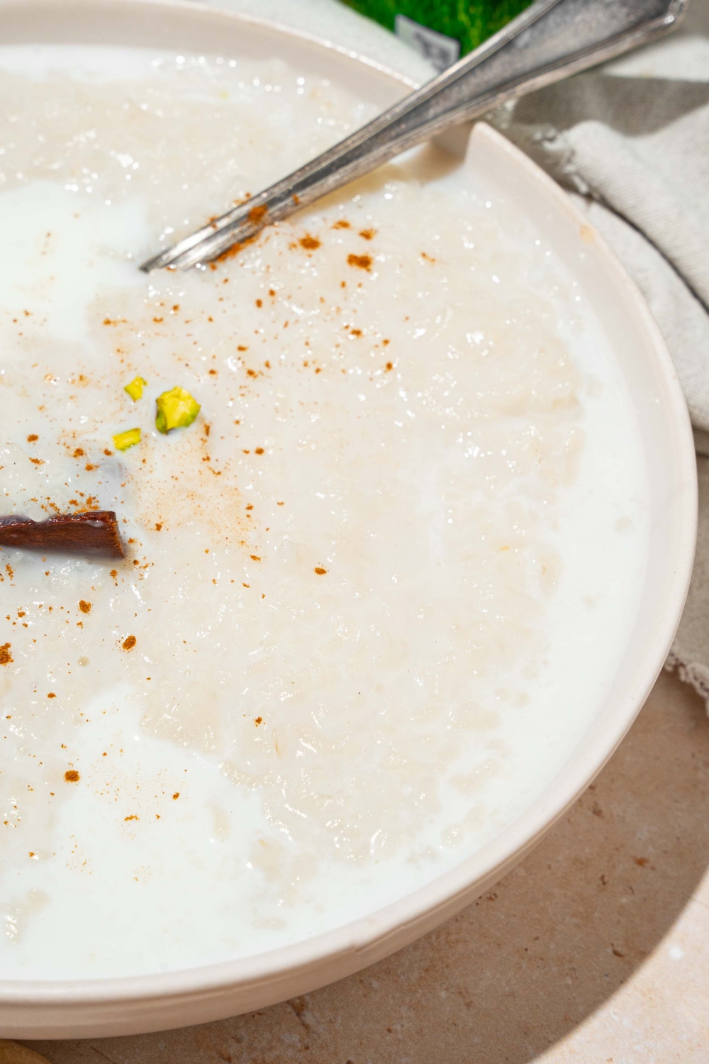 A white bowl with arroz con leche topped with a cinnamon stick and pistachios. There is a spoon in the bowl.