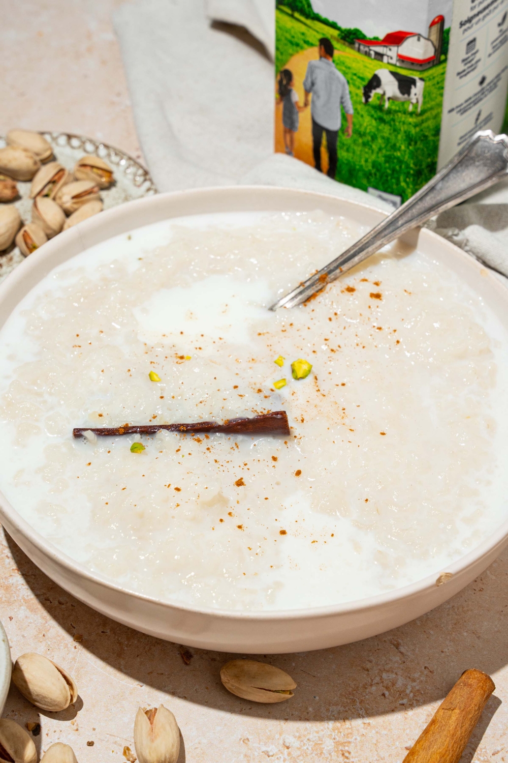 A white bowl with arroz con leche topped with a cinnamon stick and pistachios. There is a spoon in the bowl. The bowl is on a tan counter with a plate of pistachios.