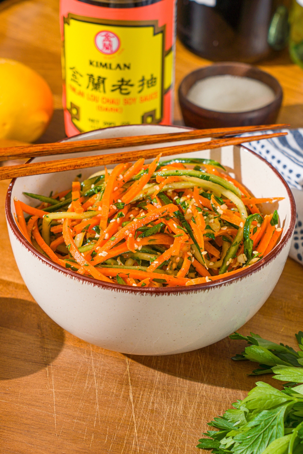 A white bowl with carrot cucumber salad tossed in dressing and garnished with sesame seeds. There is a pair of chopsticks on top of the bowl. The bowl is on a wooden board with a jar of soy sauce and small bowl of garnishes.