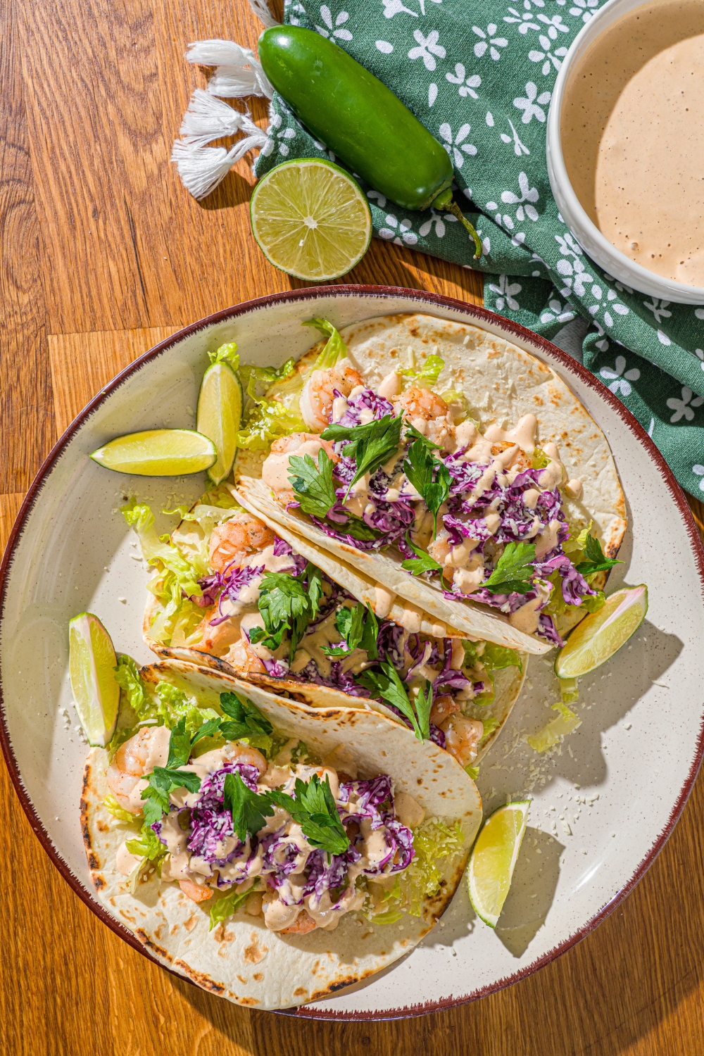 A white plate with three shrimp tacos topped with shredded cabbage, baja sauce, and fresh cilantro. The plate is on a wooden counter with lime and a bowl of baja sauce with a green cloth napkin.