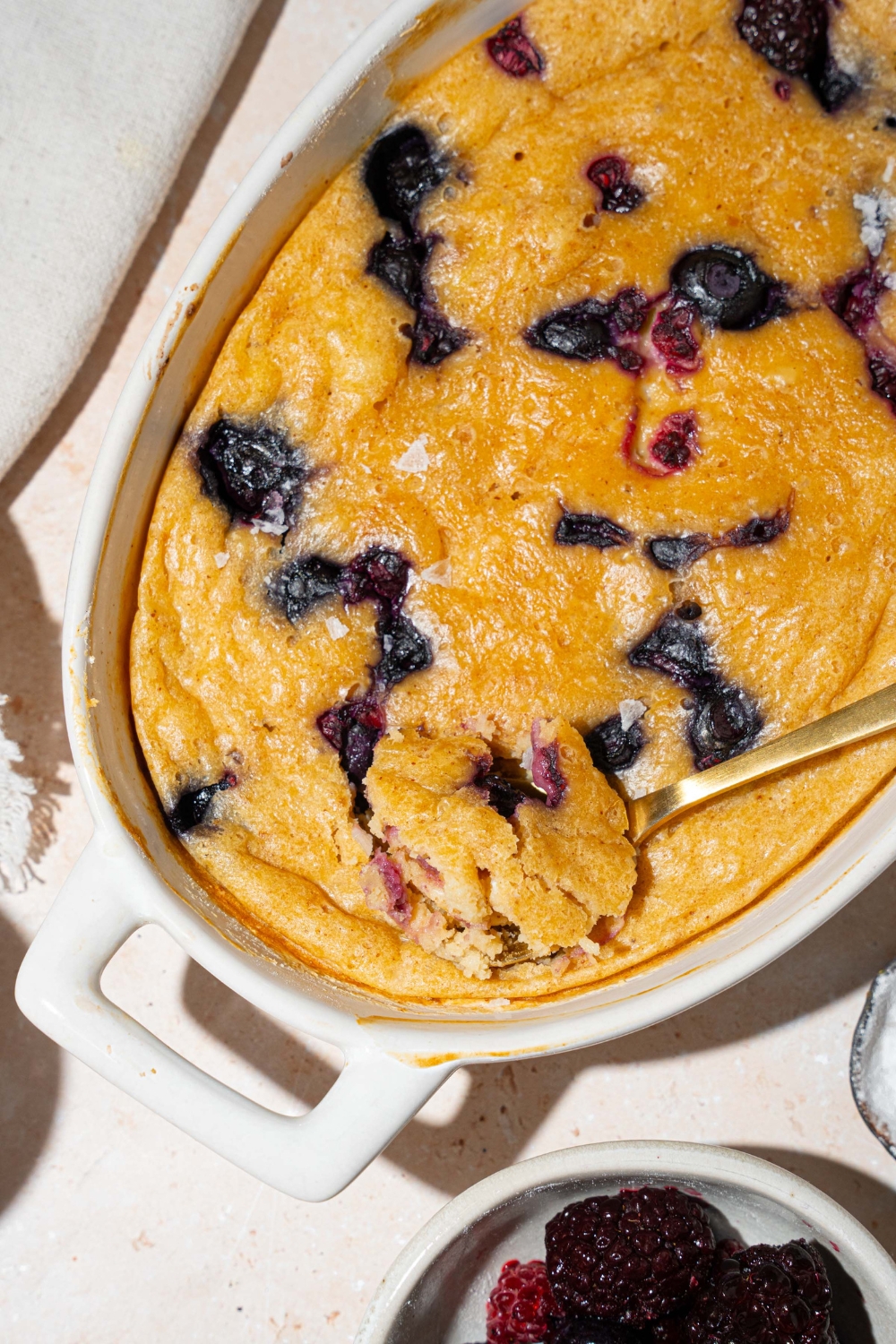 A baked protein pancake bowl with blueberries. There is a spoon taking a bite from the bowl. The bowl is on a tan counter with a white cloth napkin and small bowl of fruit.