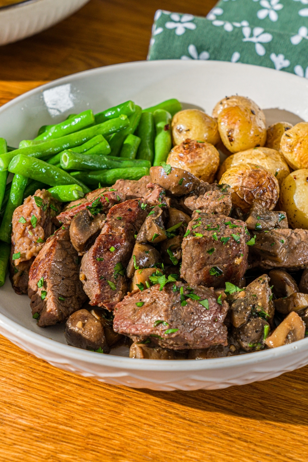 A white plate with tenderloin tips with mushrooms served with roasted potatoes and green beans. The bowl is garnished with fresh parsley. The bowl is on a wooden counter with a green cloth napkin.