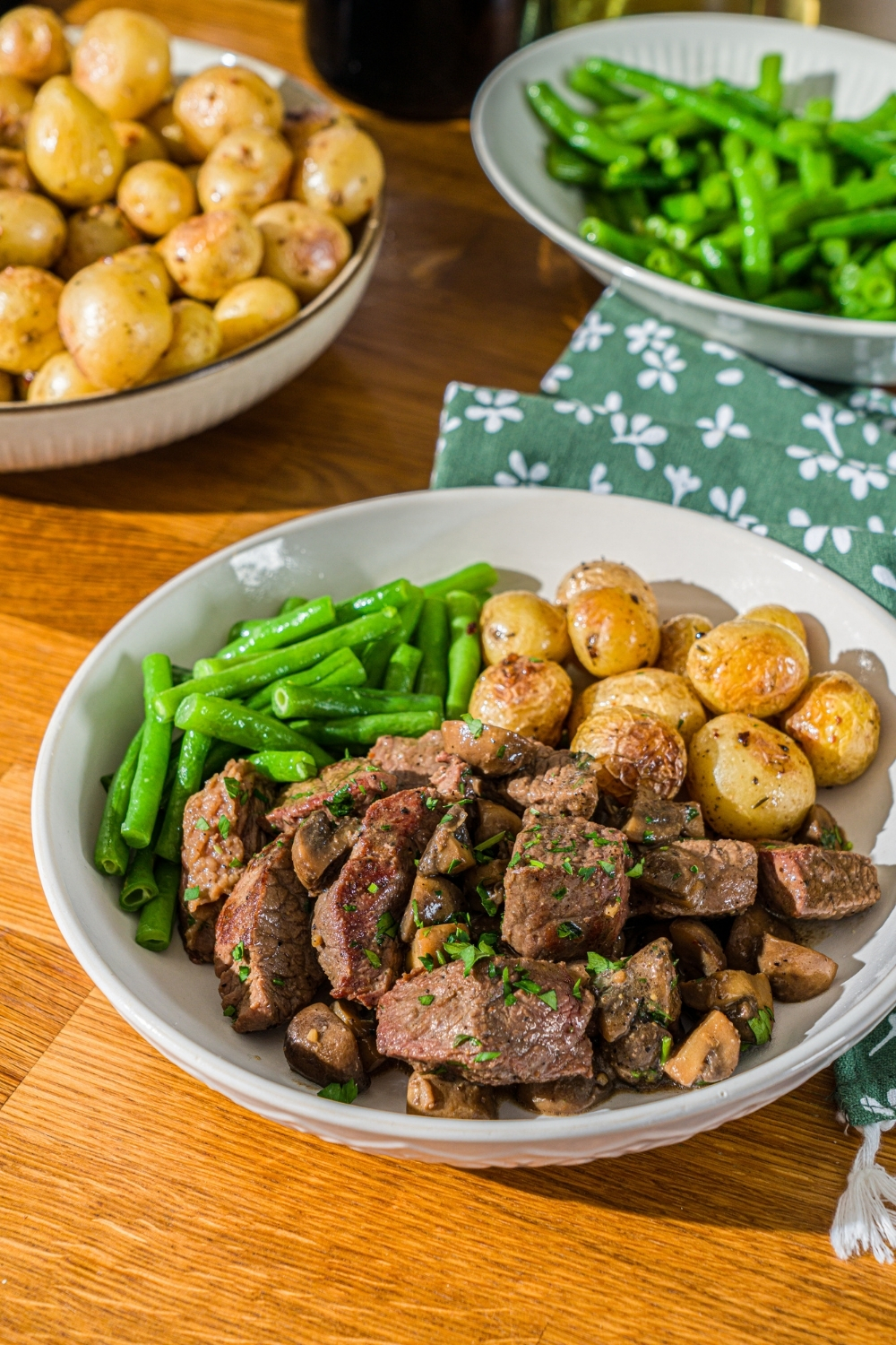 A white plate with tenderloin tips with mushrooms served with roasted potatoes and green beans. The bowl is garnished with fresh parsley. The bowl is on a wooden counter with a green cloth napkin and bowls of potatoes and green beans.