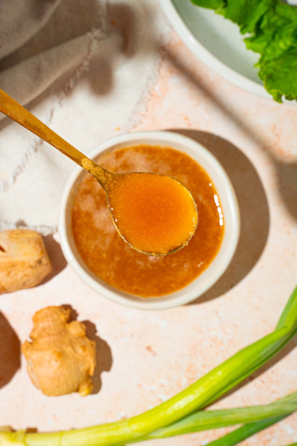 A small white bowl of hibachi ginger sauce with a spoon in the bowl. The bowl is on a tan counter with ginger, a plate with lettuce, and a white cloth napkin.