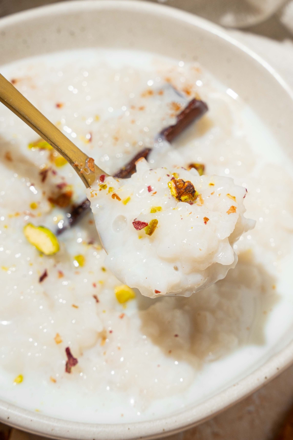 A white bowl with arroz con leche topped with a cinnamon stick and pistachios. A spoon is taking a bite from the bowl.