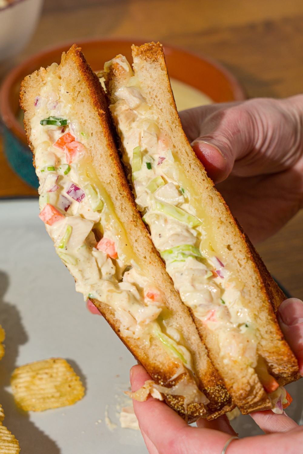 A hand holding a chicken melt sliced in half. There is a plate with chips in the background.