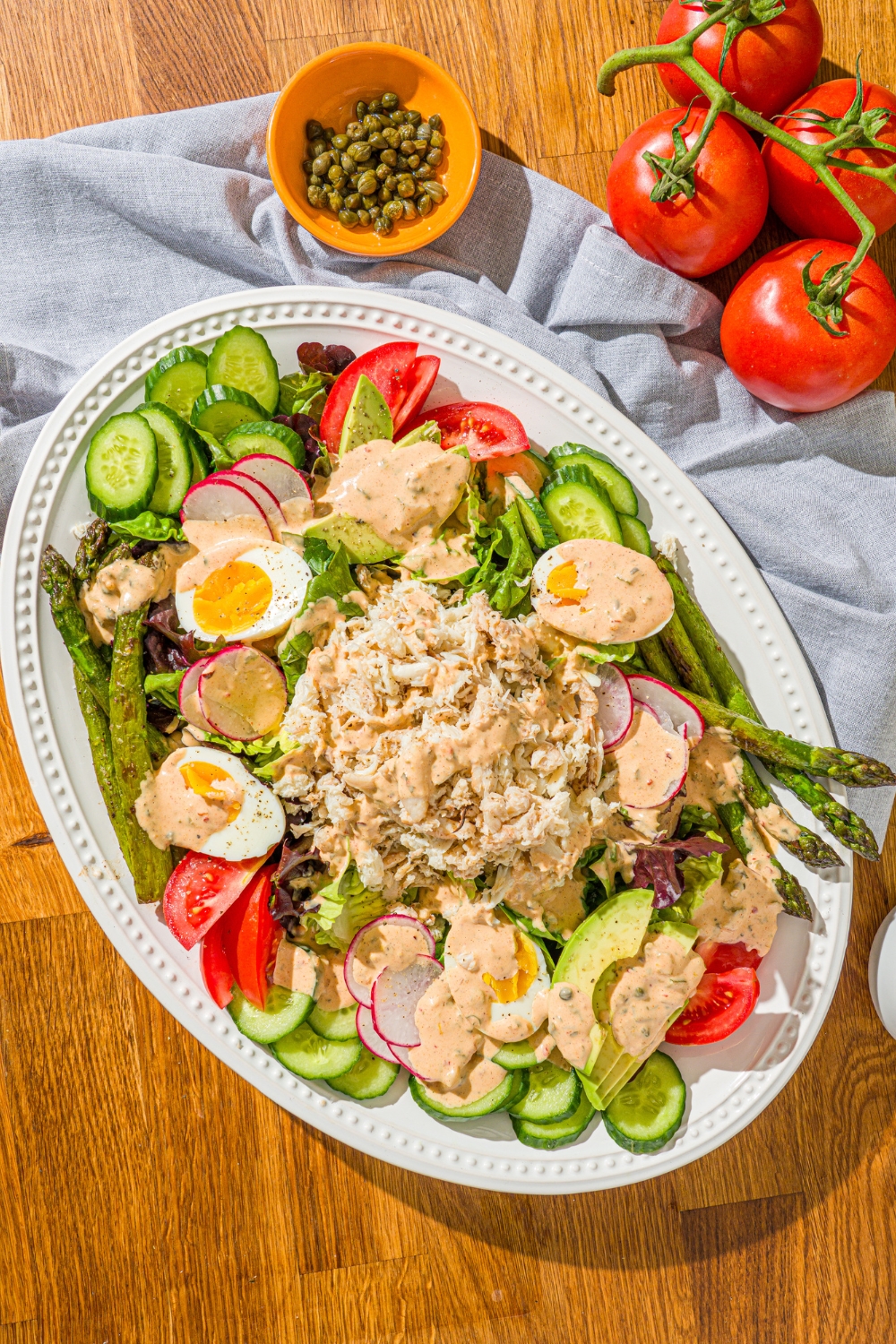An oval platter with crab Louie salad including a bed of lettuce, hard boiled egg, asparagus, sliced vegetables, and lump crab drizzled with a crab Louie salad dressing. The platter is on a wooden counter with a blue cloth napkin and a bunch of tomatoes.