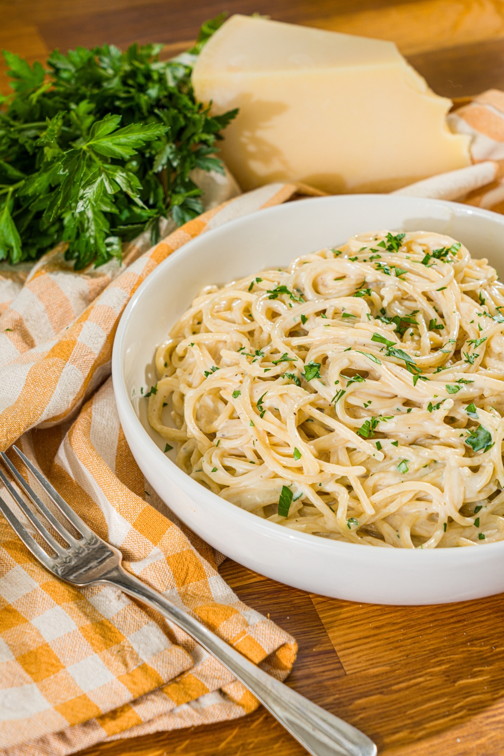 A white bowl with spaghetti tossed in a creamy garlic parmesan sauce garnished with fresh parsley. The bowl is on a wooden counter with a fork and yellow checkered napkin.