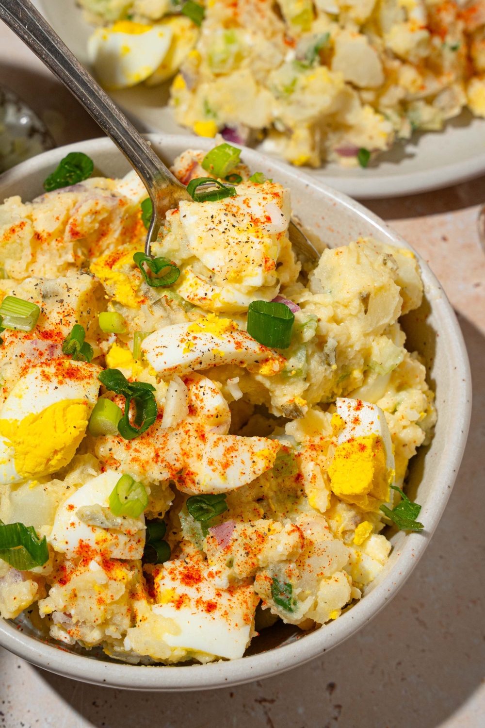 A white bowl with deviled egg potato salad garnished with sliced green egg and paprika. There is a fork taking a bite from the bowl. The bowl is on a tan counter with an additional plate of potato salad.