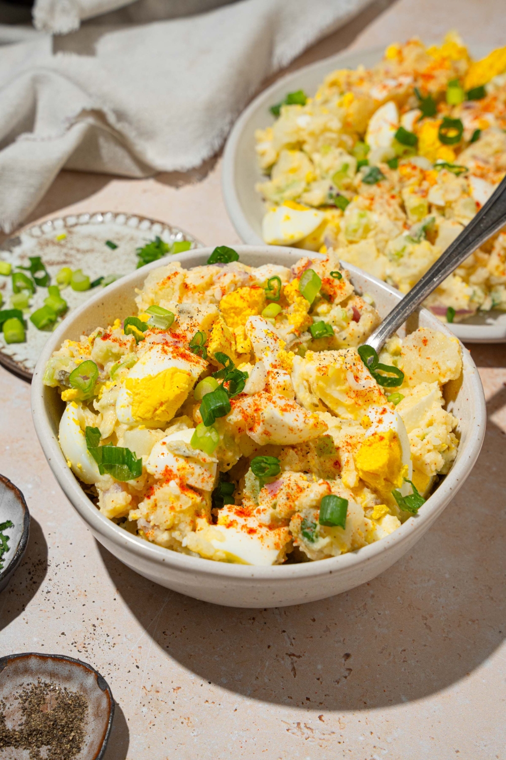 A white bowl with deviled egg potato salad garnished with sliced green egg and paprika. There is a fork in the bowl. The bowl is on a tan counter with an additional plate of potato salad.