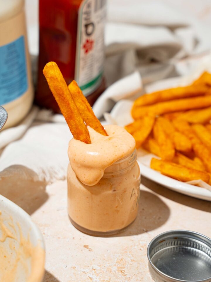 A jar of fry sauce with french fries dipped in the jar. The jar is on a tan counter with a plate of fries, ketchup, and mayo.