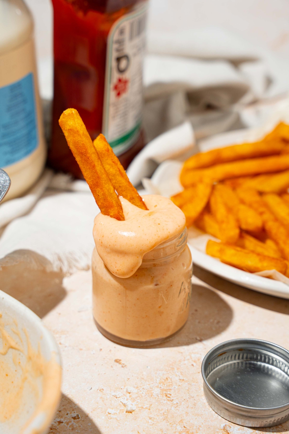 A jar of fry sauce with french fries dipped in the jar. The jar is on a tan counter with a plate of fries, ketchup, and mayo.