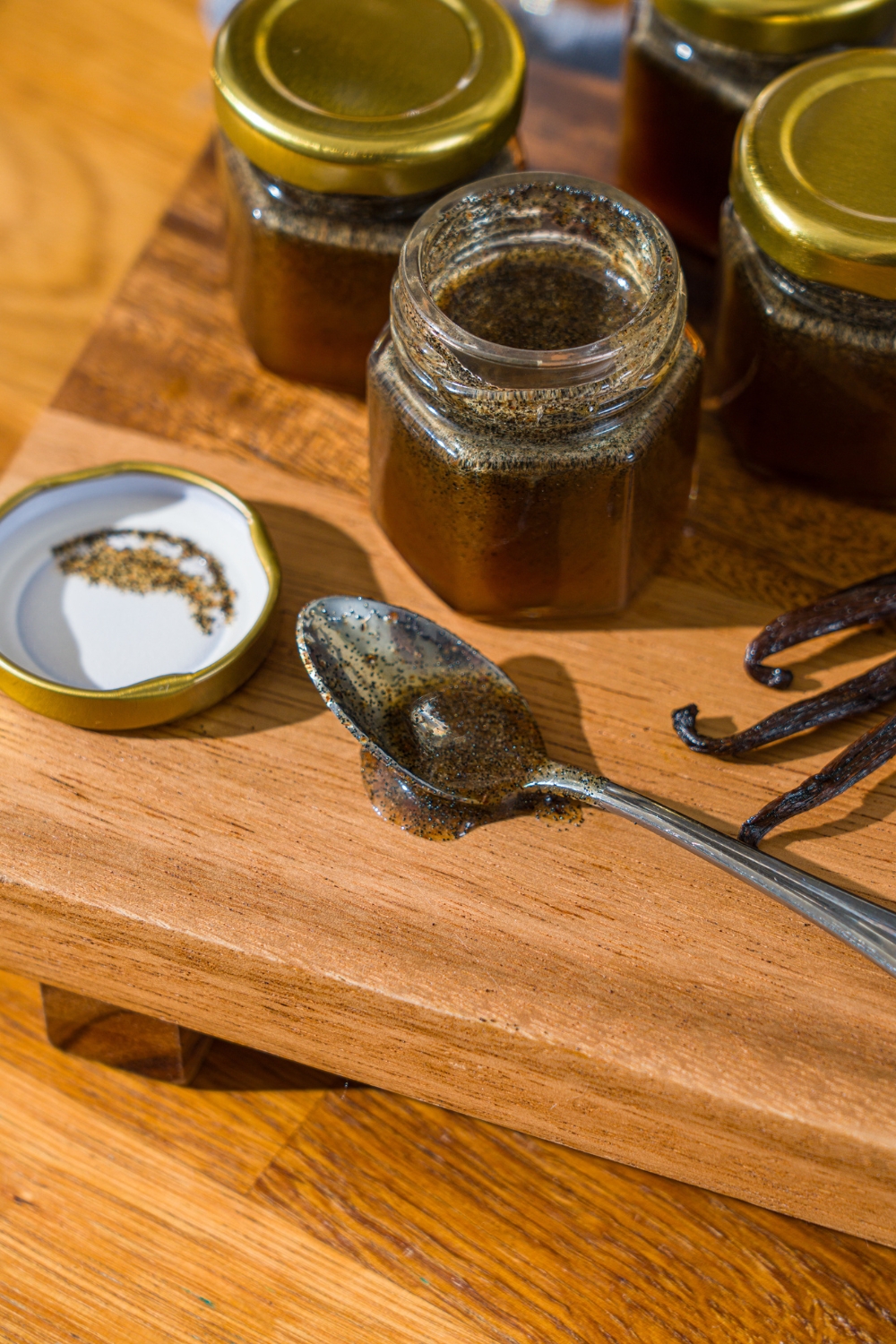 A wooden board with several jars of homemade vanilla bean paste with whole vanilla beans. There is a spoon on the board dipped in vanilla bean paste.