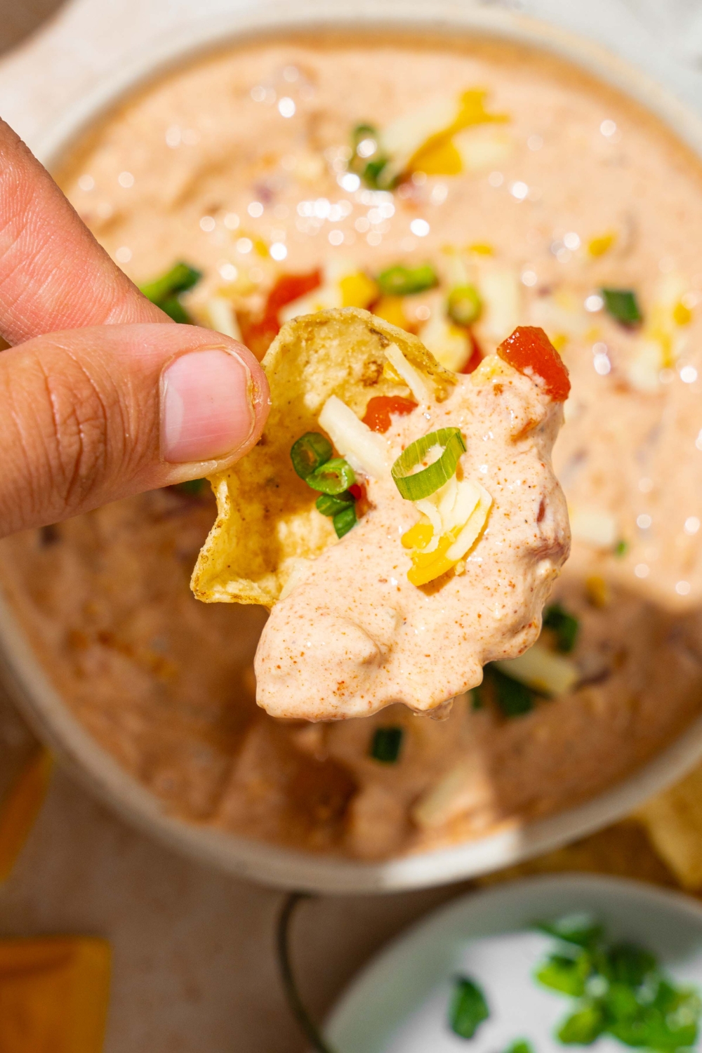 A close up of a chip dipped in poolside dip garnished with sliced green onion and shredded cheese. There is a bowl of dip blurred in the background.