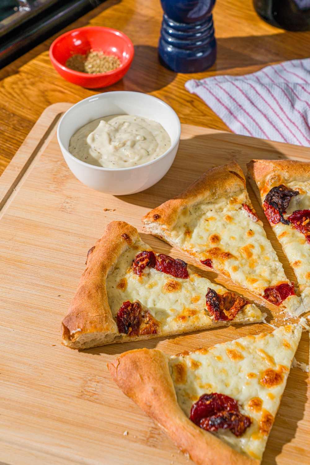 A wooden board with a sliced garlic white pizza topped with roasted peppers. There is a bowl of garlic pizza sauce on the board. The board i on a wooden counter.