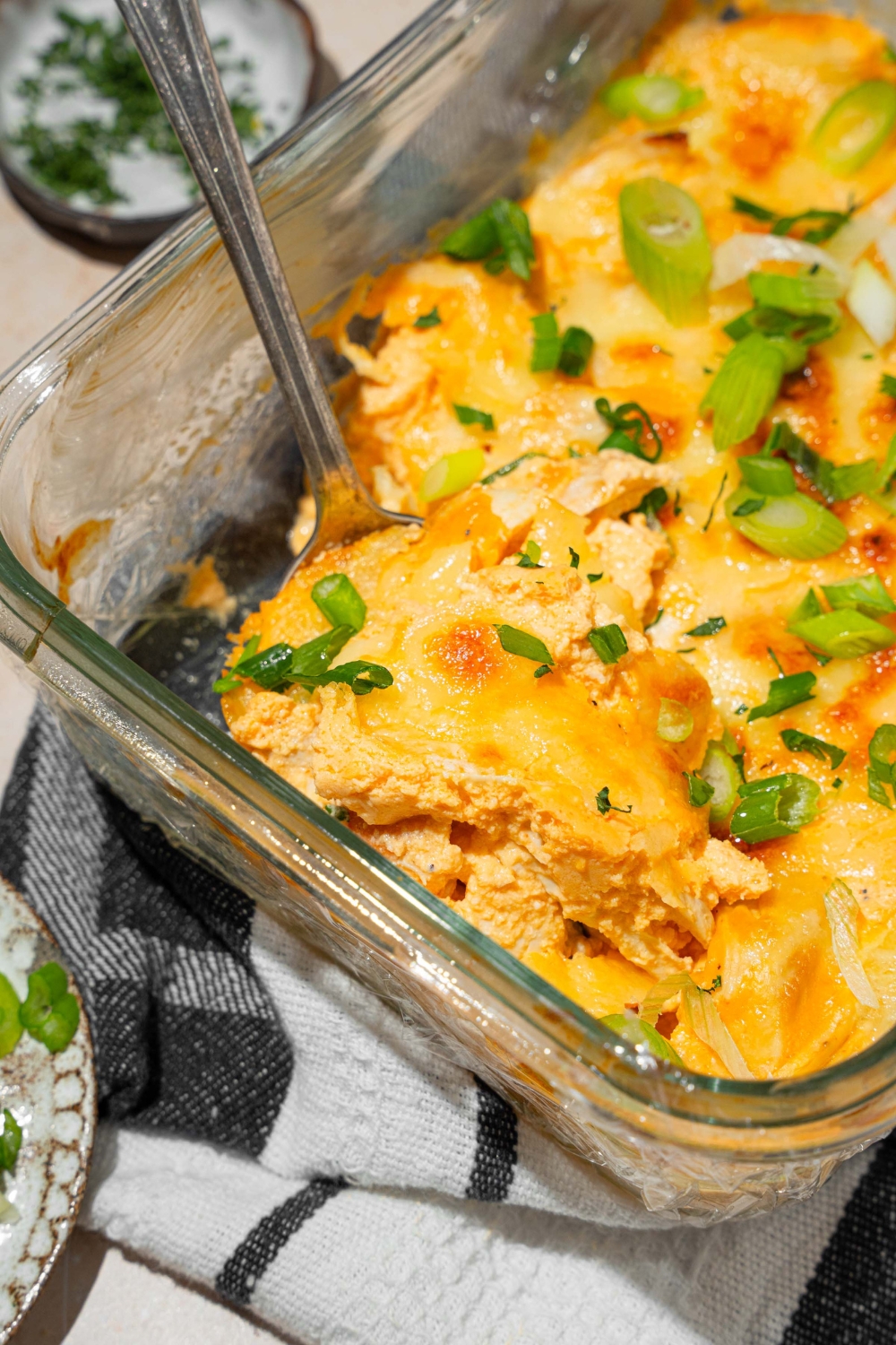 A glass baking dish with cottage cheese Buffalo chicken dip garnished with sliced green onion. A spoon is dipped in the dip. The dish is on a dish towel on a tan counter with a small plate of garnishes.