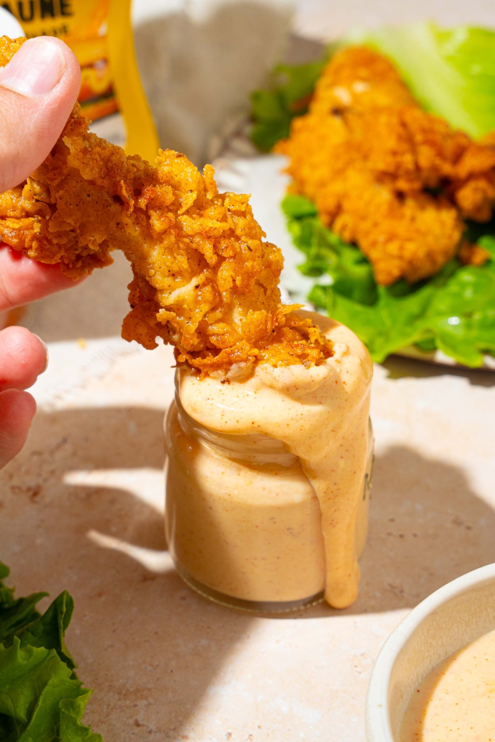 A crispy chicken strip dipping into a jar of Burger King zesty sauce. The jar is on a tan counter with a plate of chicken strips with lettuce.