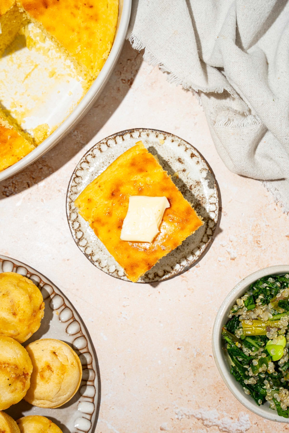 A plate with a slice of cornbread without eggs topped with a pat of butter. The plate is on a tan counter with a plate of muffins and white cloth napkin.