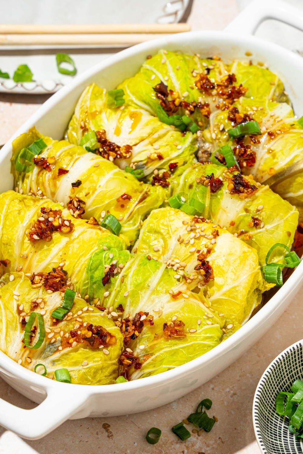 An oval baking dish with baked cabbage dumplings topped with chili oil, sesame seeds, and sliced green onion. The dish is on a tan counter with a small bowl of garnishes.