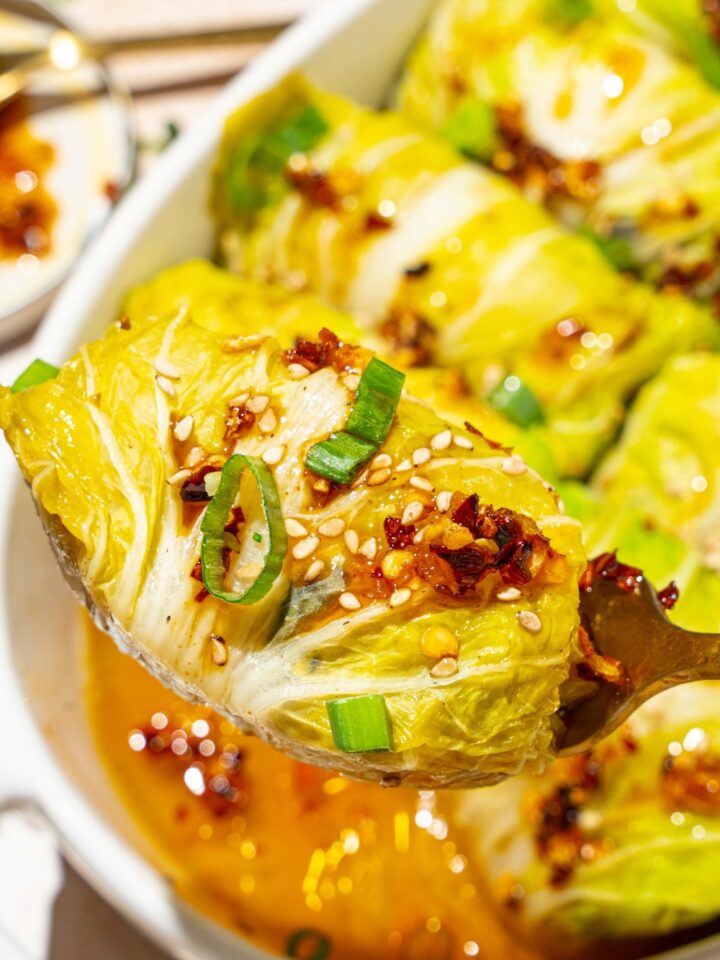 A close up of a spoon holding a cabbage dumpling topped with chili oil, sesame seeds, and sliced green onion. There is a baking dish of dumplings blurred in the background.