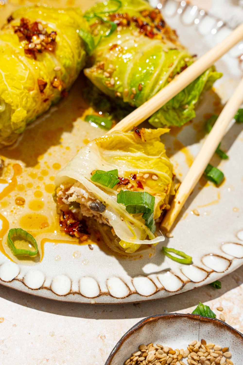 A white plate with three pieces of cabbage dumplings topped with chili oil, sesame seeds, and sliced green onion. A pair of chopsticks are taking a dumpling with a bite removed from it.