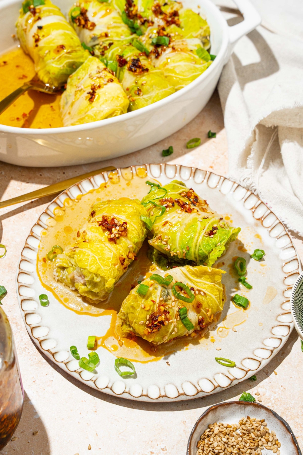 A white plate with three pieces of cabbage dumplings topped with chili oil, sesame seeds, and sliced green onion. The plate is on a tan counter with a baking dish of dumplings and small bowls of garnishes.