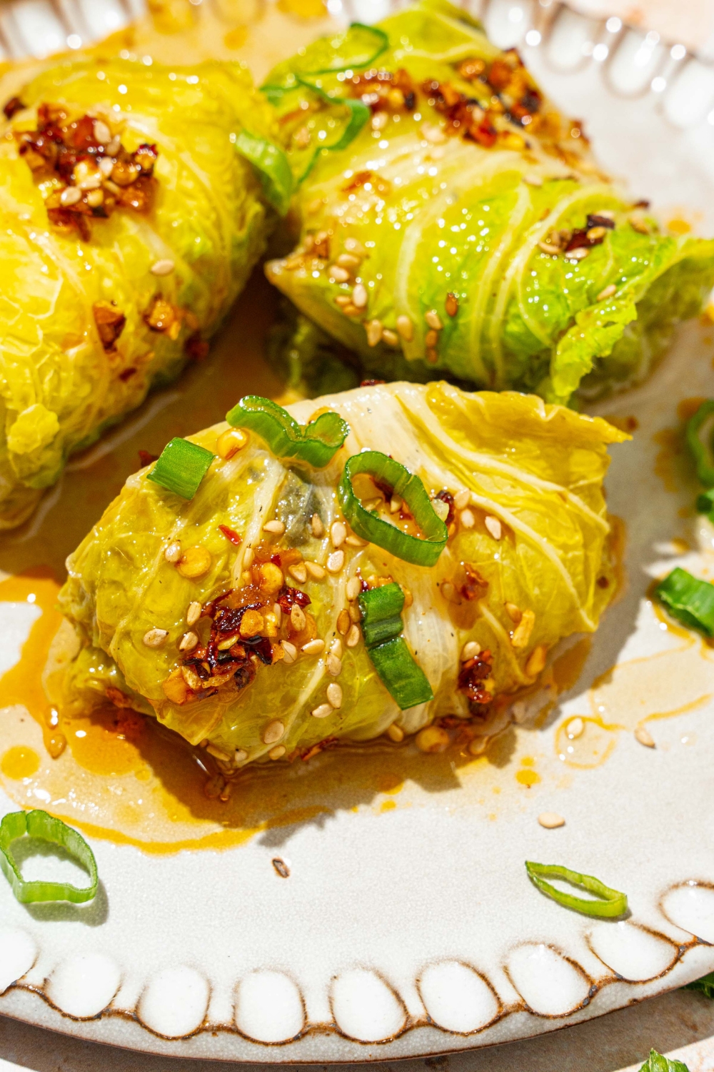 A white plate with three pieces of cabbage dumplings topped with chili oil, sesame seeds, and sliced green onion.