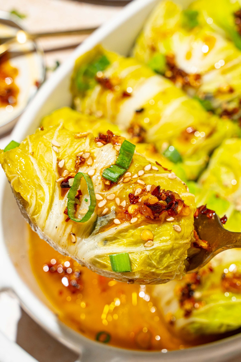 A close up of a spoon holding a cabbage dumpling topped with chili oil, sesame seeds, and sliced green onion. There is a baking dish of dumplings blurred in the background.