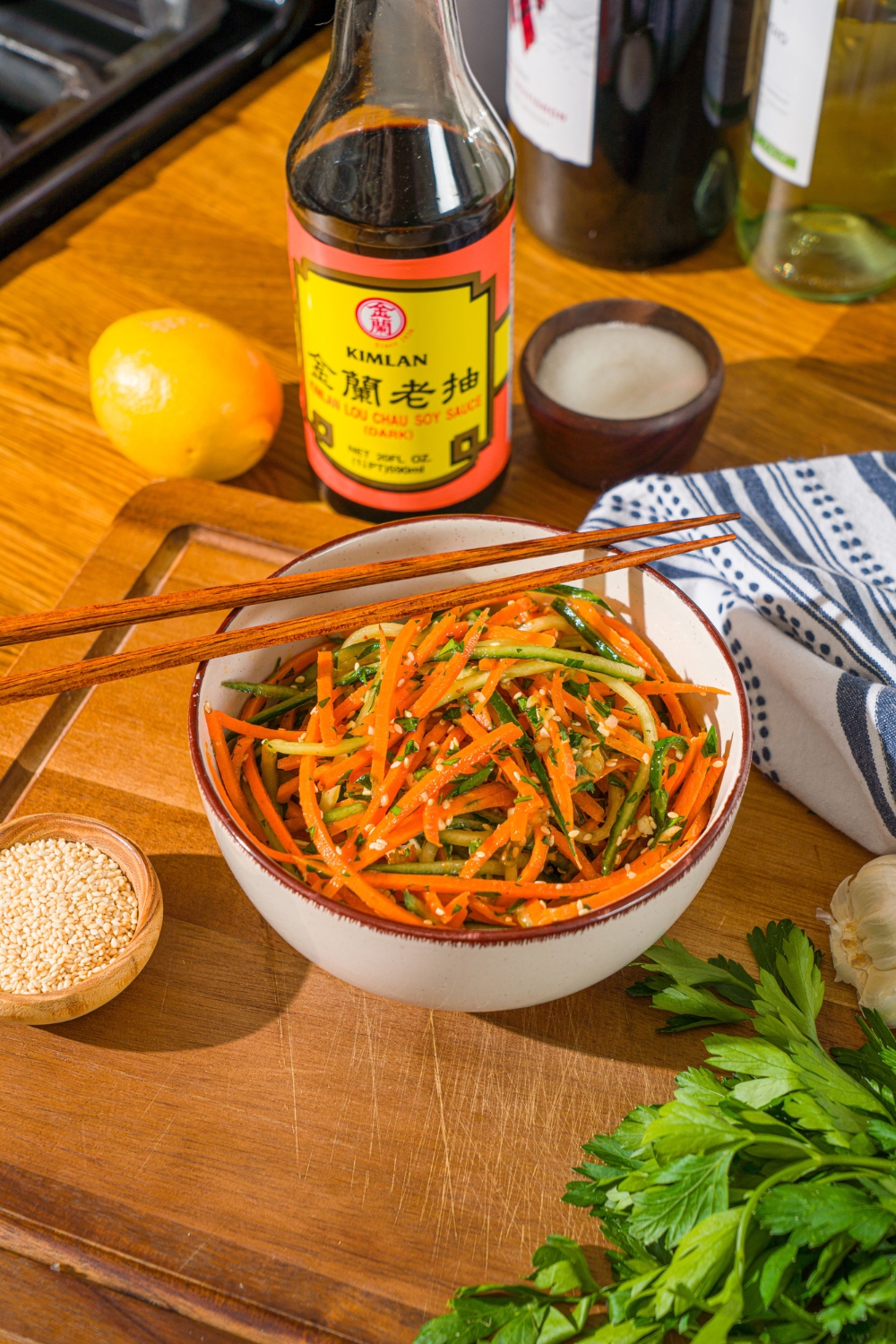 A white bowl with carrot cucumber salad tossed in dressing and garnished with sesame seeds. There is a pair of chopsticks on top of the bowl. The bowl is on a wooden board with a jar of soy sauce and small bowl of garnishes.