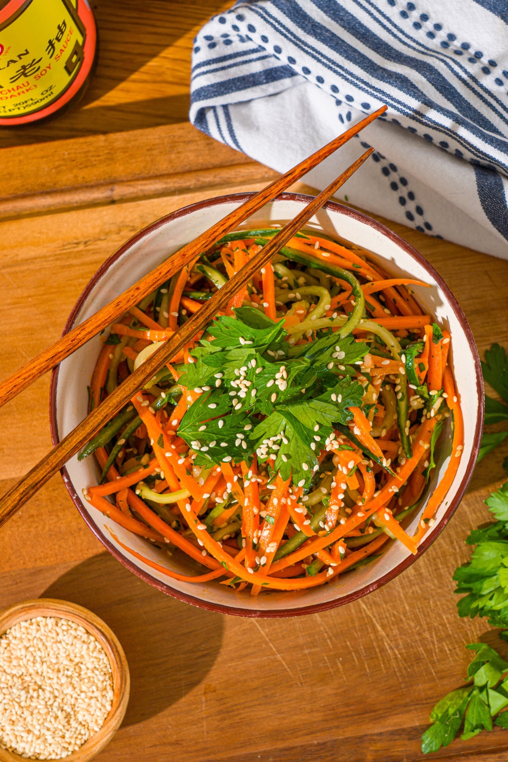 A white bowl with carrot cucumber salad tossed in dressing and garnished with fresh parsley and sesame seeds. There is a pair of chopsticks on top of the bowl. The bowl is on a wooden board with a small bowl of sesame seeds and white cloth napkin.