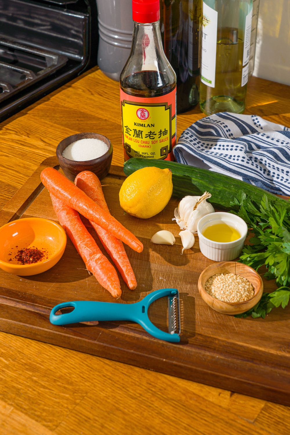 A wooden cutting board with ingredients to make carrot cucumber salad including carrots, cucumber, lemon, parsley, soy sauce, olive oil, garlic, sesame seeds, and seasonings.